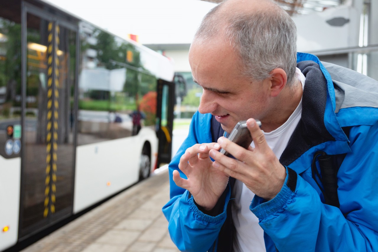 Midadult blind man, wearing casual blue clothing, holding his cellphone near his face, listening to information. He is standing at a bus station