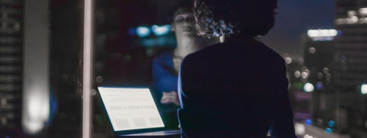 Stylishly Dressed Businesswoman Holds Laptop While Looking Out of the Window of Her Office. Late at Night Woman Doing Important Job. Window Has Big City Business District View with Many Night Lights.