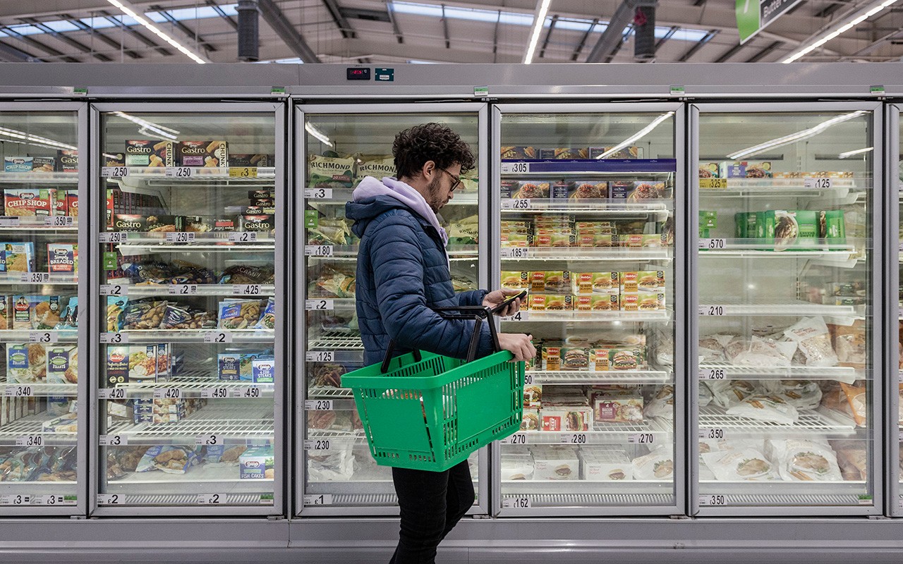 Man shopping in a supermarket while on a budget. He is looking for low prices due to inflation, standing looking at his phone in front of a row of freezers. He is living in the North East of England.