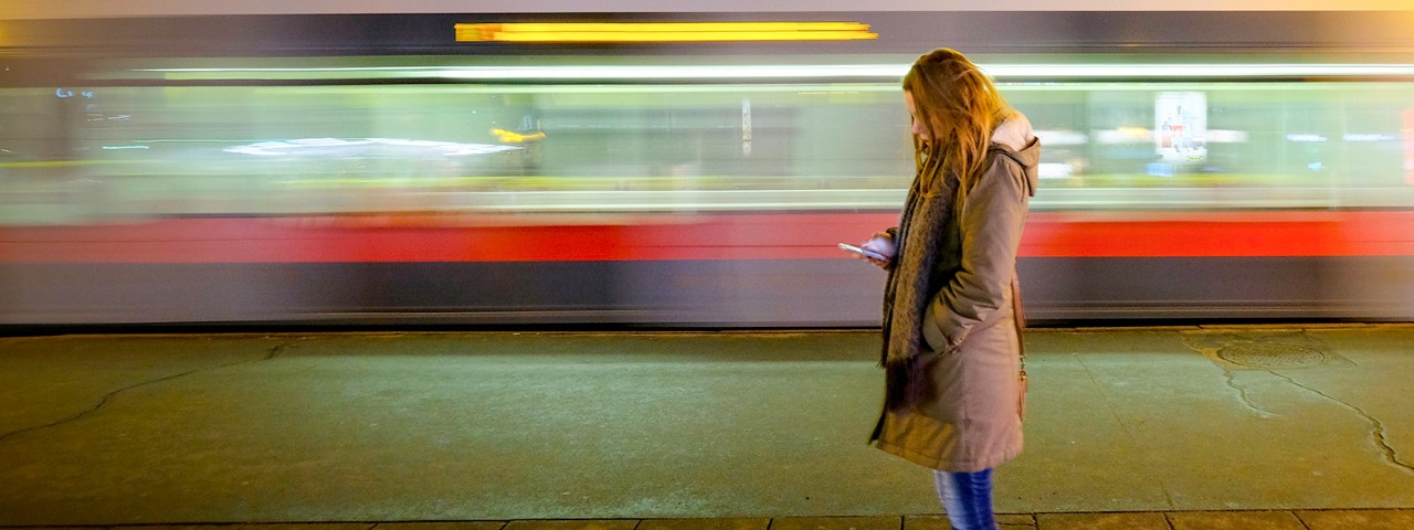 Woman standing alone on city street and checking her smartphone at night. Driving tram in background.