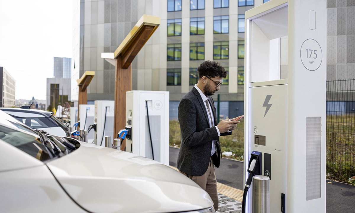 A wide-angle side view of a man in business dress using his mobile phone to access an app to be able to put his electric vehicle on charge at an electrical charging station in Newcastle City Centre in the North East of England. It is in a row of electric charging points.